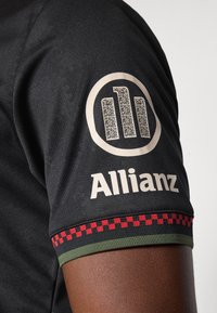 Jersey negro con un acabado texturizado. Presenta un logo con un patrón y un código QR en la manga, además de un acento a cuadros verde-rojo en el puño.