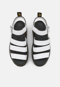 Hvit stropp sandal med svarte såler og gule sting. Har justerbar ankelrem og polstret såle. Toppvisning.