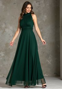 Ciemnozielona suknia maxi z wysokim dekoltem i plisowanym topem. Zwiewna spódnica ma przezroczystą warstwę wierzchnią. Widoczne czarne szpilki.