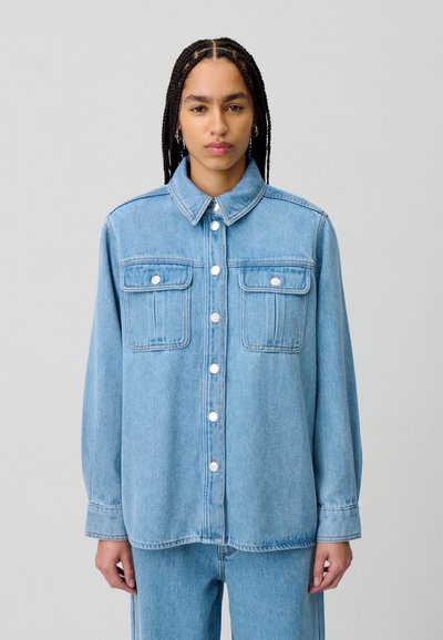 Chemise en denim bleu clair avec col, fermeture à boutons et deux poches poitrine. Présente des manches longues et un design décontracté.