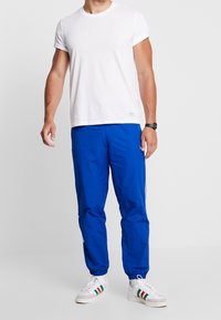 Vit t-shirt med rund hals, blå träningsbyxor med elastiska muddar och vita sneakers med gröna, röda och svarta detaljer. Enkel, avslappnad look.