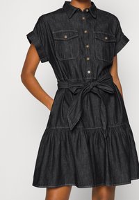 Dark denim dress with short sleeves, collar, button placket, two chest pockets, and a tied waist. Features a flared skirt with gathered detail.