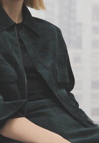 Personne portant une veste en velours côtelé vert foncé et une jupe assortie, assise avec les mains posées sur les genoux, fond clair flou.
