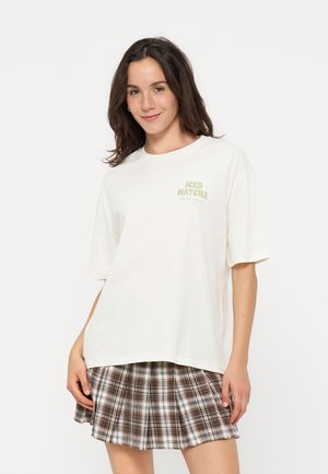 Jeune femme portant un T-shirt blanc oversize avec le texte « Iced Matcha » et une jupe plissée à carreaux marron, souriante avec la main posée sur la jupe.