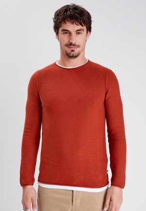Long-sleeve rust-colored shirt with textured fabric and a contrasting white round neckline, paired with light beige pants.