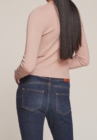 Vaaleanpunainen neule, jossa pitkät hihat ja resorireuna, yhdistetty keskikorkeavyötäröisiin tummansinisiin farkkuihin, joissa on takataskut ja ruskea nahkalappu.