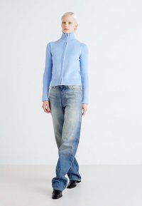 Svetlo modra zip-up jopica z ribasto teksturo in prilegajočo zasnovo, v kombinaciji s svetlo zbledelimi ohlapnimi kavbojkami in črnimi čevlji.