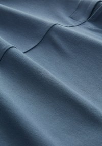 Sininen kangas, jossa on sileä, hieman teksturoitu pinta, esittelee hellävaraisia laskoksia ja saumoja, mikä viittaa kevyeseen ja hengittävään materiaaliin.