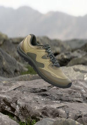 Scarpa da trekking beige e grigio scuro con suola scolpita, sospesa in aria sopra un terreno roccioso con montagne sfocate sullo sfondo.