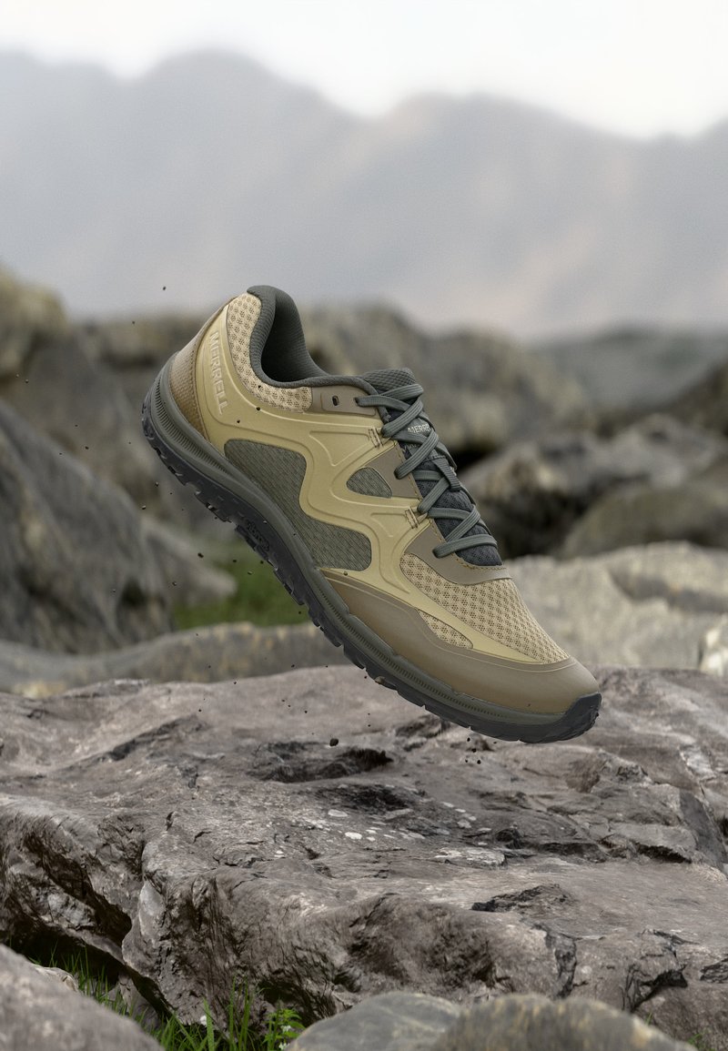 Chaussure de randonnée beige et gris foncé avec semelle crantée en l'air au-dessus d'un terrain rocheux, avec des montagnes floues en arrière-plan.