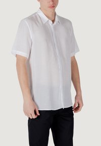 Camisa blanca de manga corta hecha de un tejido ligero, con cuello y cierre de botones en la parte frontal. Presenta una textura suave y un diseño clásico.