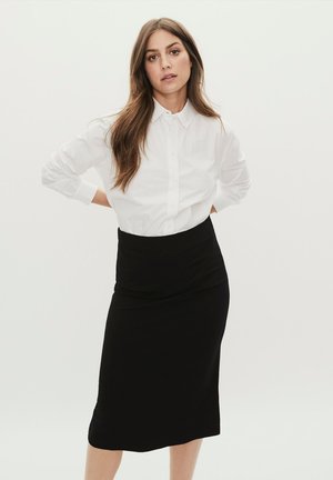 Frau mit langen braunen Haaren, die ein weißes Hemd mit Knöpfen und einen schwarzen knielangen Rock trägt, steht mit den Händen hinter dem Rücken.