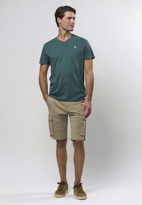 Grønn v-hals t-skjorte, beige cargoshorts og brune joggesko. T-skjorten har en liten logo, shortsen har sidelommer og en avslappet passform.