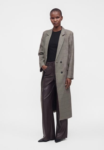 Femme portant un long manteau à carreaux par-dessus une chemise noire et un pantalon en cuir large et sombre, avec des chaussures noires pointues, debout avec une main dans la poche.