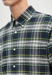 Knappskjorte med rutemønster i marineblått, grønt og gult med hvite detaljer. Laget av glatt stoff med brystlomme.