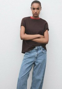 Camiseta de manga corta marrón oscuro con cuello rojo, combinada con unos jeans de pierna ancha de color azul claro, ligeramente desgastados, frente a un fondo neutro.