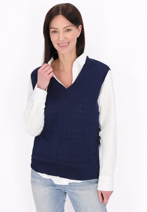 Kvinde smiler iført en hvid skjorte under en mørkeblå strikket ærmeløs sweater med lyseblå jeans på en hvid baggrund.