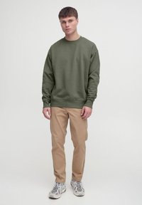 Olivzaļš džemperis ar apaļu kaklu, brīvs piegriezums un ribotām aprocēm, kombinēts ar haki biksēm un gaišām sporta kurpēm.