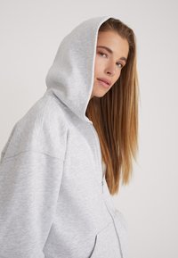 Grijze zip-up hoodie met een grote capuchon, zachte textuur en een losse pasvorm. Heeft een voorkantzak en zichtbare stiksels.