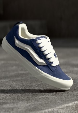 Blå og hvid lav sneaker med hvide snørebånd og gummisål, der svæver over en tekstureret grå overflade.