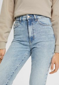 Jean en denim bleu clair taille haute avec une texture délavée, design à cinq poches et fermeture à bouton en métal. Accentué par des coutures dorées.