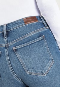 Os jeans de cintura alta em denim azul apresentam um patch de couro castanho com a marca "Hollister" e dois bolsos traseiros com costura contrastante.