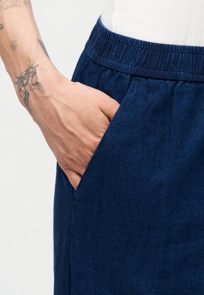 Χέρι με τατουάζ που ακουμπά μέσα στην ελαστική τσέπη της μέσης σκούρων μπλε παντελονιών σε λευκό φόντο.