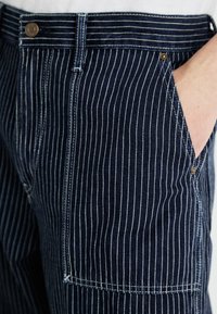Marineblau gestreifte Jeanshose. Zu den Merkmalen gehören vertikale weiße Streifen, eine gerade Passform und sichtbare Nähte mit Metallknopf.