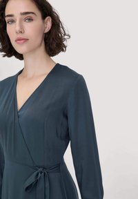 Teal-värinen kietaisumekko pitkillä hihoilla, syvällä V-kaula-aukolla ja itse solmittavalla vyöllä vyötäröllä, valmistettu sileästä, teksturoidusta kankaasta.