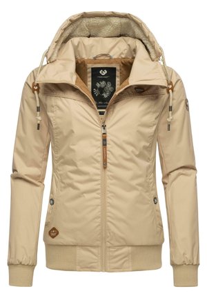 Veste beige à capuche avec doublure en polaire, fermeture à glissière sur le devant, capuche avec cordon de serrage, poignets et ourlet côtelés, et plusieurs patchs de marque ainsi que des tirettes de fermeture éclair.