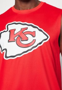 Débardeur de sport rouge avec un large contour blanc mettant en avant un logo "KC" noir et rouge. Tissu lisse et encolure ronde.