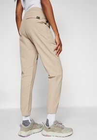 Pantalons de jogging tapered beiges avec des poignets élastiques, une poche arrière et une étiquette de marque. Associés à des baskets vert clair et des chaussettes blanches.