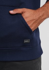 Sudadera azul marino con un bolsillo tipo canguro y dobladillo acanalado; presenta una pequeña etiqueta de logo en blanco y negro de O'Neill en el lado.