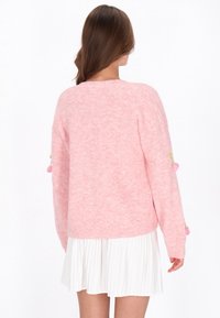 Rosa stickad tröja med ribbad textur, med färgglada pom-pom detaljer på ärmarna, i kombination med en veckad vit kjol.