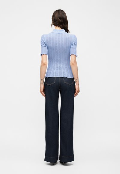 Γυναίκα που στέκεται πλάτη, φοράει ανοιχτό μπλε κοντομάνικη ριμπ πλεκτή μπλούζα και σκούρα φαρδιά τζιν με γυρισμένα τελειώματα.