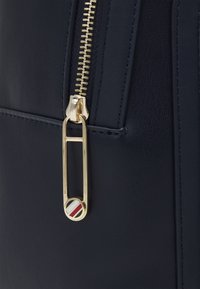 Záběr na zlatý zip na tmavě modré kožené tašce s prodlouženým zipovým táhlem ve tvaru bezpečnostní špendlíku, které má červené, bílé a modré pruhy.