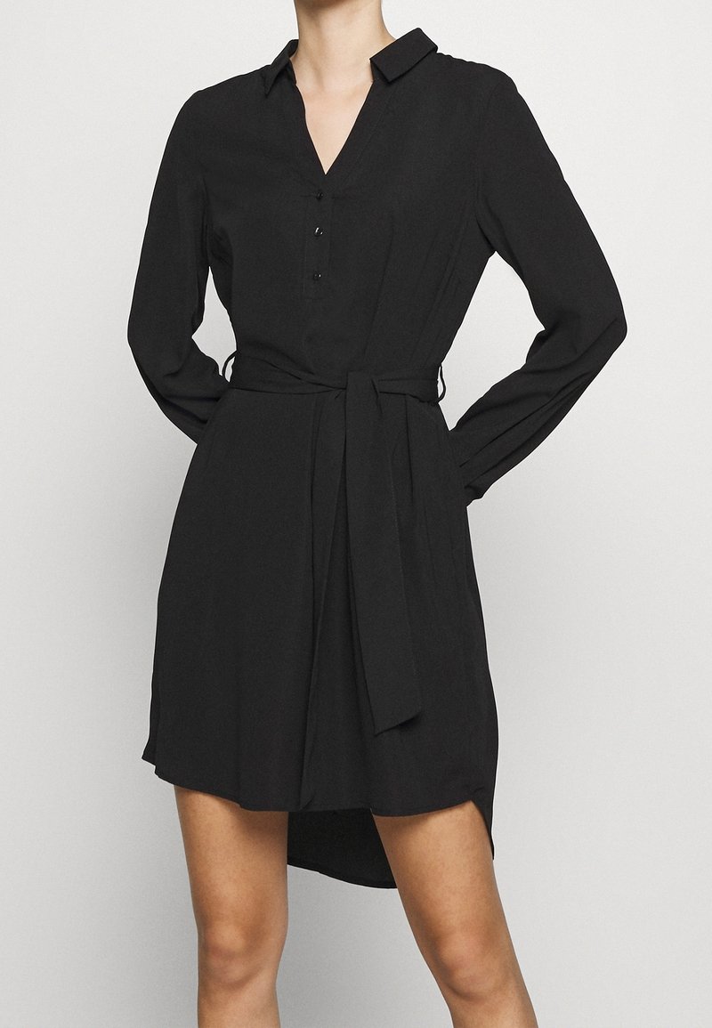 Robe chemise noire en tissu léger, dotée d'un col en V boutonné, de manches longues et d'une ceinture nouée à la taille avec un ourlet asymétrique.