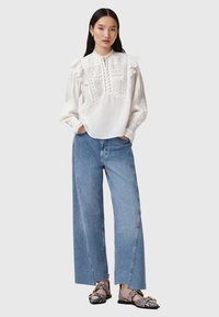 Blusa bianca con maniche a volant e cuciture dettagliate, abbinata a jeans a gamba larga di un blu chiaro. Footwear presenta un motivo testurizzato.