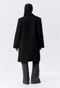 Person with braided hair wearing a black knee-length coat and gray wide-leg pants standing facing away against a plain light background.