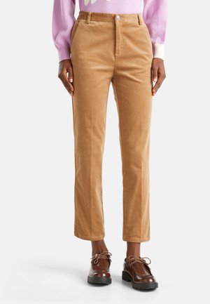 Persoon in lichtbruine corduroy broek, bruine leren veterschoenen en een lavendelkleurige trui met witte manchetten, staande tegen een witte achtergrond.