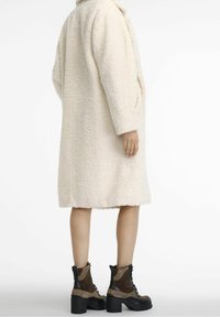 Długi płaszcz w kremowym kolorze, wykonany z puszystej tkaniny, o luźnym kroju i bocznych kieszeniach. Zestawiony z brązowo-czarnymi botkami na obcasie.