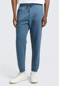 Niebieskie, zwężane spodnie dresowe z elastycznym pasem i sznurkiem, wyposażone w boczne kieszenie i ściągacze u dołu nogawek, wykonane z miękkiego materiału.