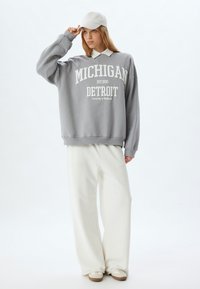 Übergröße grauer Sweatshirt mit "MICHIGAN DETROIT" Druck, kombiniert mit weit geschnittenen weißen Hosen und einer hellgrauen Mütze. Weiches Material und lockere Passform.