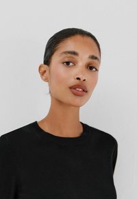 Maglione nero a maglia con collo rotondo, texture liscia e silhouette aderente. Il modello ha capelli lisci e trucco naturale, mostrando un'espressione neutra.