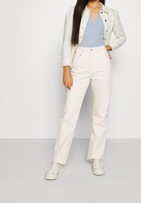 Chaqueta de mezclilla blanca sobre una blusa de canalé azul claro, combinada con pantalones blancos de tiro alto y zapatillas ligeras. Diseño simple, silueta ajustada.
