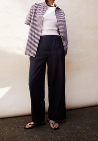 Osoba v fialové kostkované košili s krátkým rukávem přes bílém tílku, v námořnických kalhotách s širokými nohavicemi a v černých sandálech na pásku, stojí na chodníku.