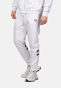 Witte sportbroek van lichtgewicht materiaal, met een elastische tailleband, marineblauwe accentstrepen en een logo op de linker bovenbeen.