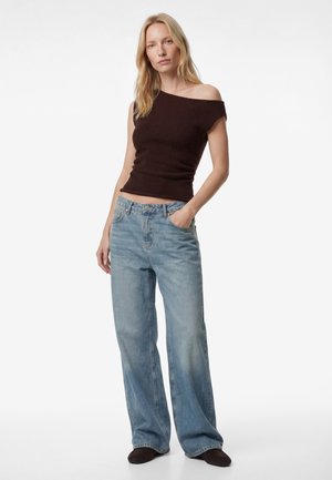 Vrouw met blond haar draagt een donkerbruine top met blote schouders, lichtblauwe wijde jeans en donkere schoenen, met één hand in de zak.