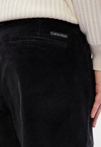 Μαύρο παντελόνι από κορντόρι με κάθετες ρίγες, διαθέτει πίσω τσέπη και ετικέτα με το λογότυπο Calvin Klein. Η υφή φαίνεται μαλακή και ανθεκτική.