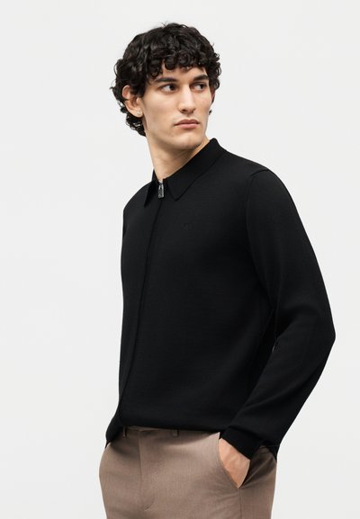 Άνδρας με σγουρά μαλλιά που φοράει μαύρο φούτερ με φερμουάρ και μπεζ παντελόνι, κοιτώντας προς τα πλάγια με το ένα χέρι στην τσέπη.